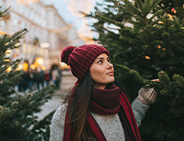 A young woman is standing between two trees in a Christmas tree lot.