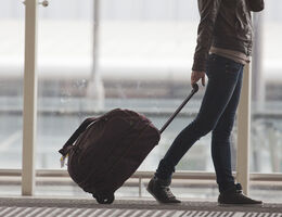  A person, seen from below the shoulders, pulls a small wheelie bag in an airport terminal.
