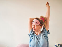 A woman stretches in bed with a sleep mask pushed up to her forehead.
