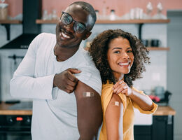 Two people stand back-to-back, each pulling their shirt sleeve up to show an adhesive bandage on their upper arm.
